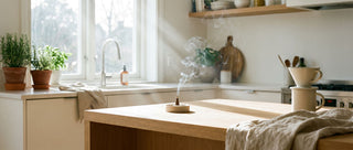 A bright, airy kitchen scene — aromatic oud incense cone burning on a matte ceramic holder on the counter, subtle wisps of smoke rising. Natural light. Clean, modern aesthetic. Lifestyle photography.